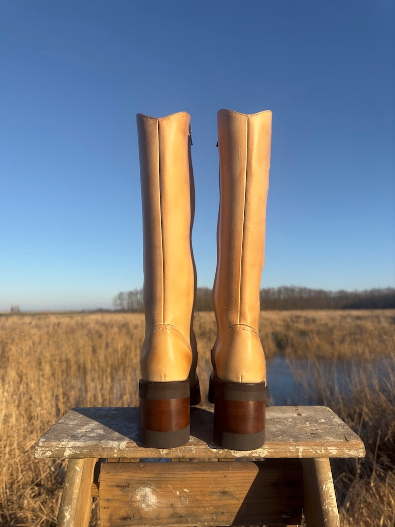 May include: A pair of tall, light brown leather boots with a chunky heel. The boots have a zipper on the inside of each boot and are sitting on a wooden stool.