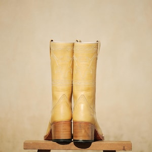 Puede incluir: Un par de botas de vaquero de color amarillo claro con costuras decorativas y un tac&oacute;n de bloque de madera. Las botas se exhiben sobre un peque&ntilde;o taburete de madera sobre un fondo neutro.