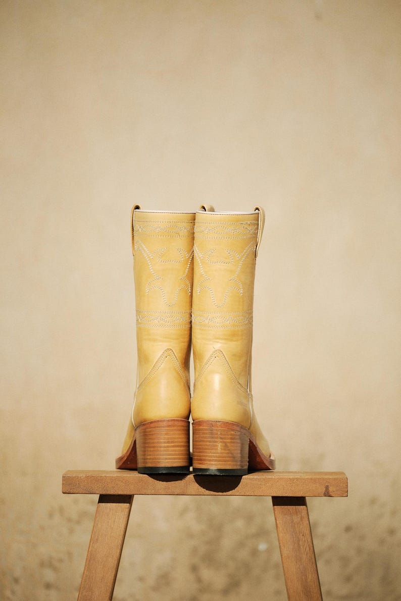 Puede incluir: Un par de botas de vaquero de color amarillo claro con costuras decorativas y un tac&oacute;n de bloque de madera. Las botas se exhiben sobre un peque&ntilde;o taburete de madera sobre un fondo neutro.