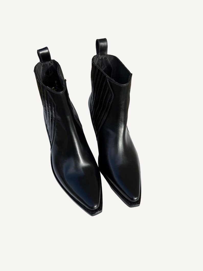 May include: A pair of black leather ankle boots with pointed toes. The boots have a pull tab at the back and a subtle, textured design on the sides. The boots are made of shiny, smooth leather.