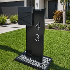 May include: A matte black metal mailbox post featuring a rectangular mailbox unit with geometric lines and the silver house numbers 4 and 3 mounted vertically on the front face. The structure is set within a rectangular base filled with dark gray stones.