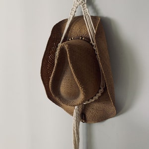 May include: A brown straw cowboy hat hanging from a white macrame plant hanger with wooden beads.
