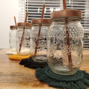 May include: Four clear glass jars with lids and straws. The jars are filled with water and have a textured surface. The jars are sitting on colourful round coasters.