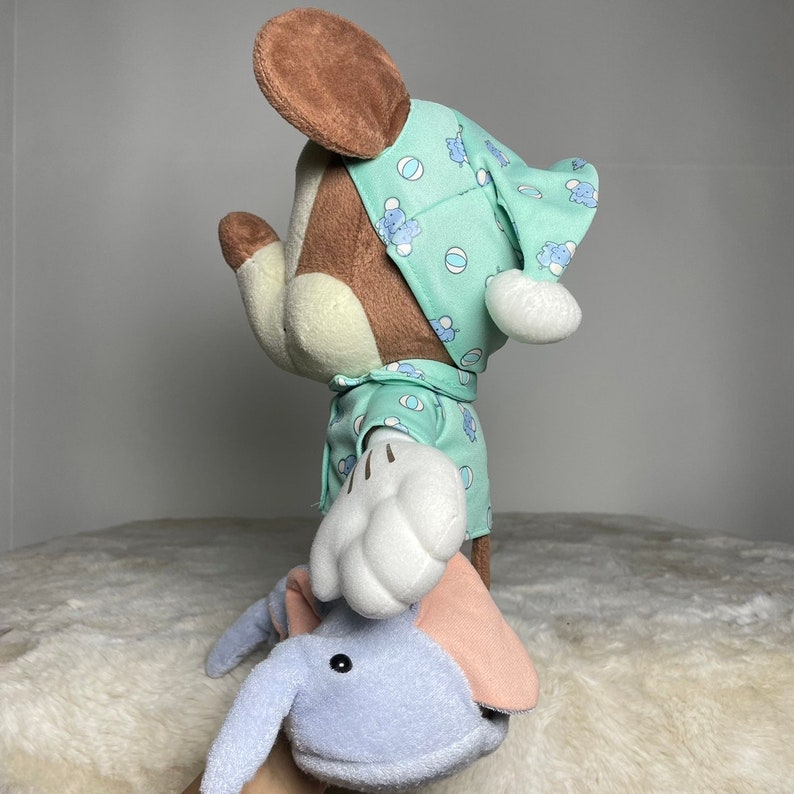Może przedstawiać: Pluszowa zabawka Mickey Mouse ubrana w zieloną i białą piżamę w wzory z dopasowaną czapką. Zabawka trzyma niebieskiego pluszowego słonia.