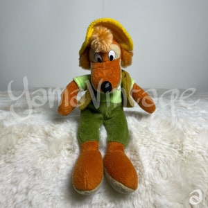 May include: A plush toy fox wearing a yellow hat and green overalls. The fox has brown fur, a black nose, and large brown eyes.