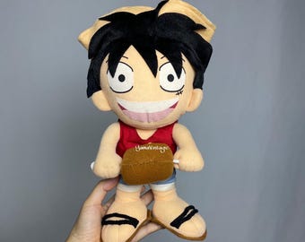 May include: A plush doll of a cartoon character with black hair, a straw hat, and a red shirt. The doll has a large smile and a scar under its left eye. The doll is holding a brown bag. The doll is wearing sandals.