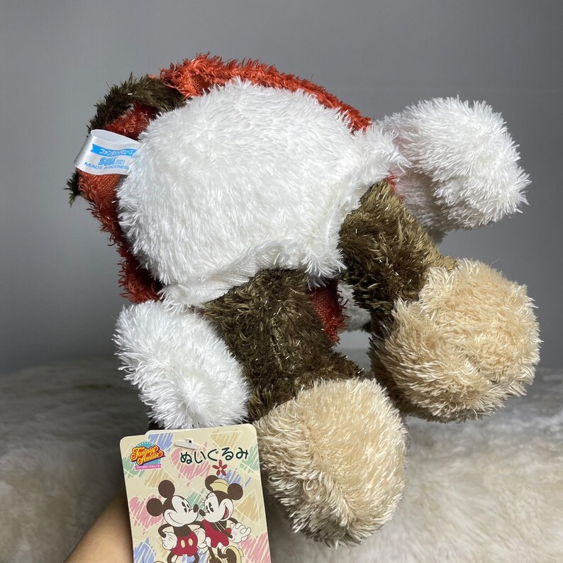 May include: A plush toy of Mickey Mouse and Minnie Mouse in a brown, white and tan colour scheme. The toy is sitting on a white surface. The tag on the toy reads "あいにくるみ" in Japanese.