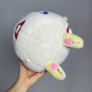 May include: White plush toy shaped like a ball with red, blue, and yellow accents. The toy has two small yellow ears with pink accents.