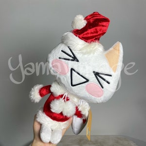 May include: White plush cat wearing a red Santa hat and a red and white Santa suit. The cat has a white fur trim around its face and a pink nose. The cat is holding a white gift bag with a gold tag.