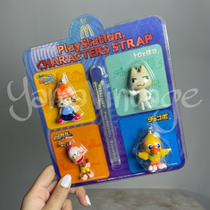 Könnte beinhalten: Ein Set mit vier PlayStation-Charakter-Anhängern, die ein gelbes Küken, eine weiße Katze, einen Affen und einen Hund zeigen. Die Anhänger sind auf einer bunten Karte mit dem Text "PlayStation Characters Strap" und "Event 2001" verpackt.