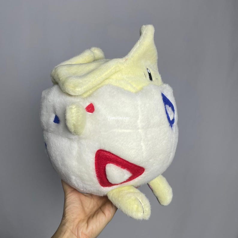 May include: A white and yellow plush toy of a Pokemon character. The toy is shaped like an egg with a yellow head and a red and blue design on the body.
