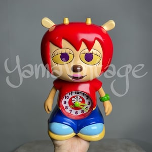 May include: Red and yellow cartoon alarm clock with a clock face and a flower design. The clock is shaped like a creature with a red head, yellow ears, and blue pants.