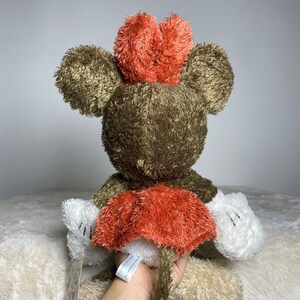 May include: A plush brown and white Mickey Mouse toy with a red bow. The toy is sitting on a white surface.