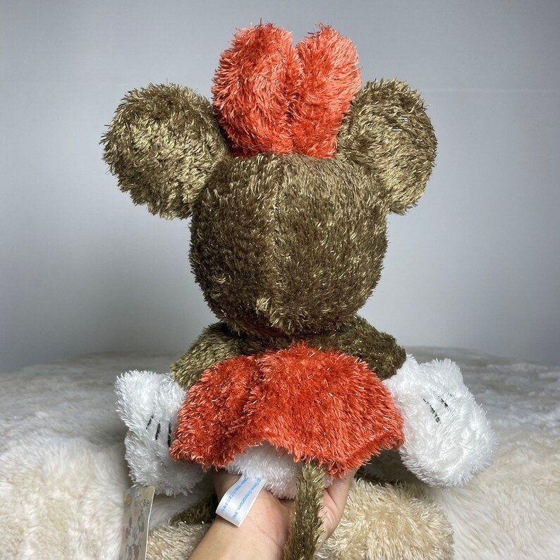 May include: A plush brown and white Mickey Mouse toy with a red bow. The toy is sitting on a white surface.