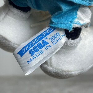 K&ouml;nnte beinhalten: Wei&szlig;es Pl&uuml;schtier mit einem blau-wei&szlig;en Outfit. Das Spielzeug hat ein Etikett mit der Aufschrift "SEGA 2000 MADE IN CHINA" in blauer Schrift auf wei&szlig;em Hintergrund.