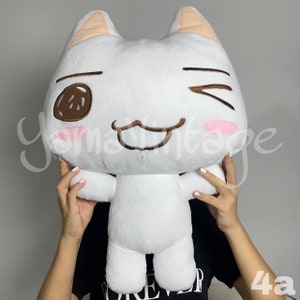 Pode incluir: Brinquedo de pelúcia de gato branco com um olho marrom, um bigode marrom e um blush rosa. O gato está piscando e tem uma expressão feliz.