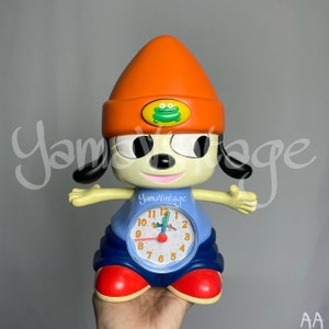 May include: A blue and orange alarm clock shaped like a cartoon dog wearing a hat. The clock face has a green frog on it and says "YamaVintage".