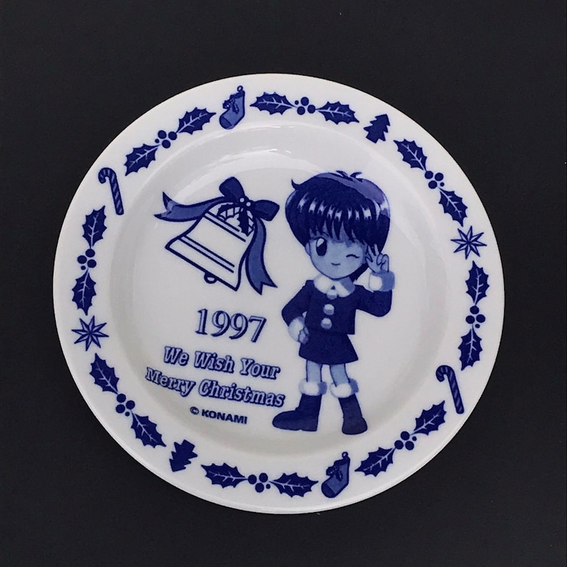 Op de afbeelding: Een wit keramiek bord met een blauwe en witte rand met een ontwerp van hulst en bessen. In het midden van het bord staat een cartoonmeisje met blauw haar, een blauwe jurk en witte laarzen. Het bord heeft ook een blauwe bel met een witte strik, een snoepstok en de tekst "1997 We Wish Your Merry Christmas &copy;KONAMI".