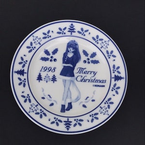 Op de afbeelding: Een wit keramiek bord met een blauw en wit ontwerp met een cartoonmeisje in een blauwe jurk en de tekst "Merry Christmas 1998 &copy;KONAMI".