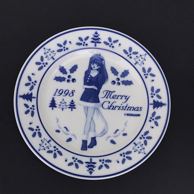 Op de afbeelding: Een wit keramiek bord met een blauw en wit ontwerp met een cartoonmeisje in een blauwe jurk en de tekst "Merry Christmas 1998 &copy;KONAMI".