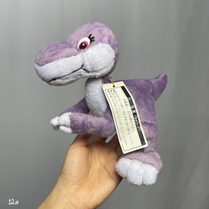 Puede incluir: Un peluche de dinosaurio morado con detalles blancos en la cara, los pies y la barriga. El dinosaurio tiene ojos rojos y una etiqueta adjunta. El juguete es suave y tierno, perfecto para niños.