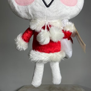 May include: White plush toy wearing a red Santa suit with white fur trim and white pom-poms. The toy is holding a white bag with a red ribbon.