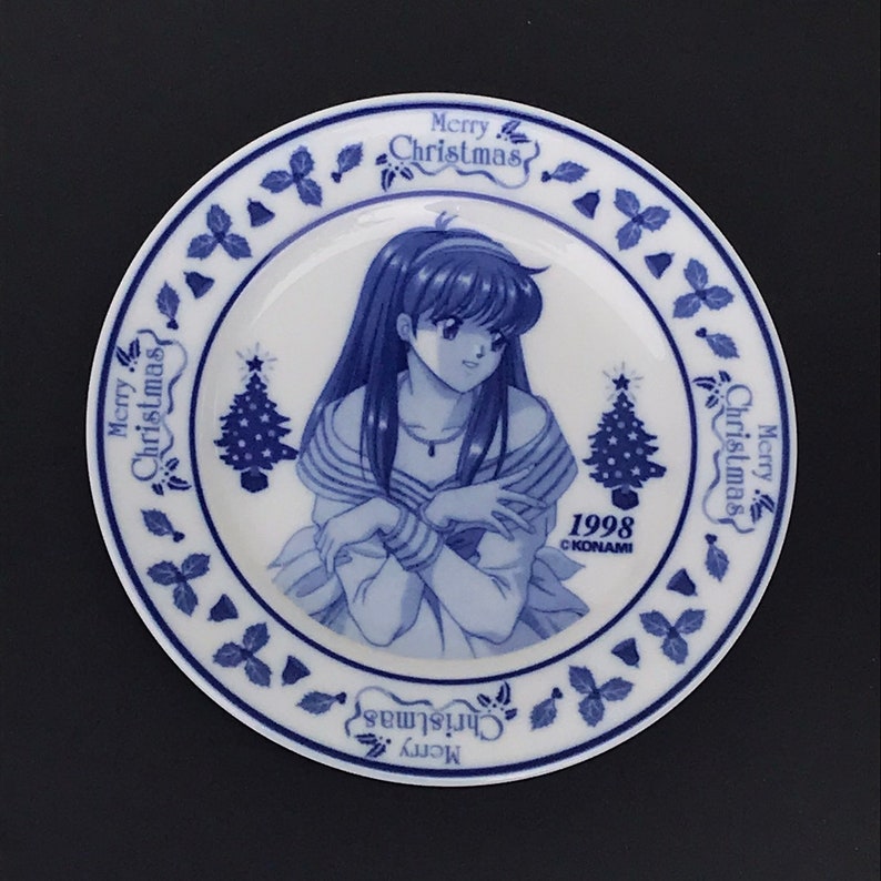 Op de afbeelding: Een blauw en wit keramiek bord met een kerstthema. Het heeft een vrouw met lang donker haar, een kerstboom en de tekst "Merry Christmas" en "1998 &copy;KONAMI".