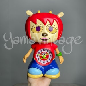May include: Red and white cartoon alarm clock with a clock face that reads "12" and "3". The clock is shaped like a character with large eyes, a red nose, and a pink mouth. The character is wearing a red shirt and blue pants.
