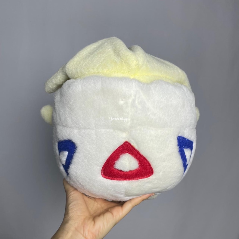 May include: White plush toy with a yellow top and red and blue triangular designs. The toy is shaped like a round head with a small mouth.