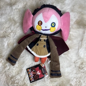 May include: A plush doll with a pink face, large pink ears, and a black and red head covering. The doll has a brown coat, brown arms, and a red cape. A tag with Japanese text is visible.
