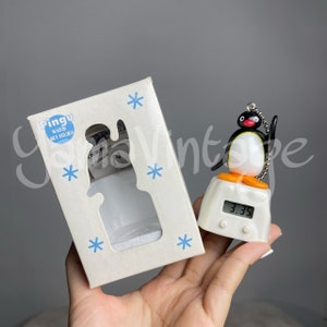 May include: A white box with a Pingu watch key holder. The box has a snowflake design and the text "Pingu Watch Key Holder". A black and white Pingu character is on a white base with a digital clock that reads 3:35.