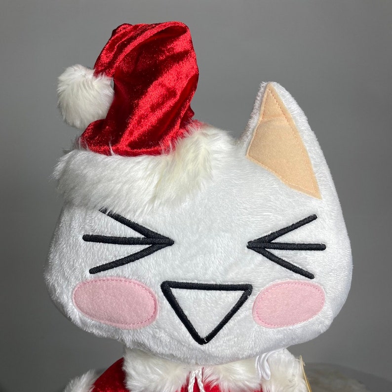 May include: White plush cat wearing a red velvet Santa hat with a white pom-pom. The cat has a smiling face with black eyes and a pink nose.