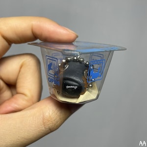 May include: A small black and brown dog keychain with a silver chain. The keychain is in a clear plastic container with the word "OPEN" printed on it.