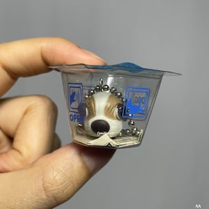 May include: A small, brown and white dog keychain with large eyes. The keychain is in a clear plastic container with a blue and white label that says "Open" and "Magg".