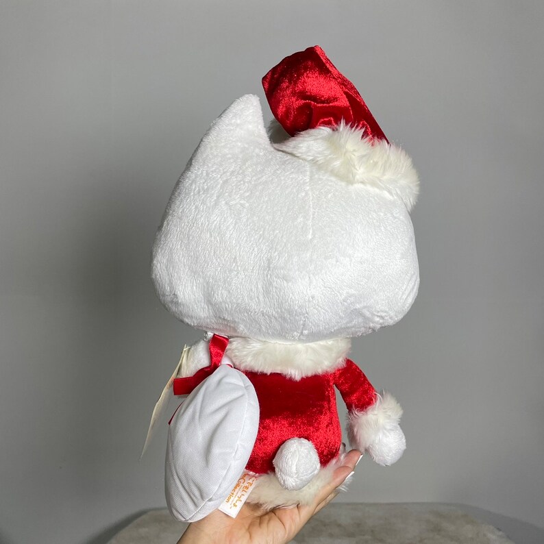 May include: White plush toy wearing a red and white Santa hat and a red velvet suit. The toy is holding a white plush gift.
