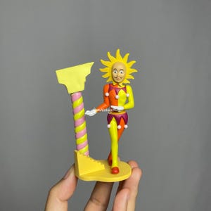 May include: A whimsical figurine of a sun-faced jester in a colorful outfit. The jester is wearing a yellow sun-shaped headdress, a harlequin-style outfit in orange, yellow, and purple, and holding a yellow structure with a pink and yellow striped pole.