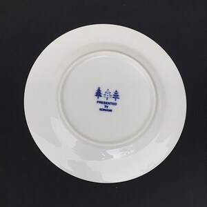 Op de afbeelding: Een wit keramiek bord met een blauw-wit logo met de tekst "PRESENTED by KONAMI" met drie gestileerde dennenbomen boven de tekst.