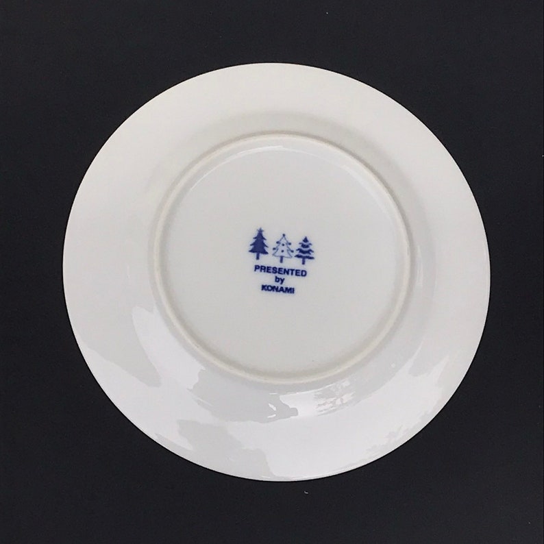 Op de afbeelding: Een wit keramiek bord met een blauw-wit logo met de tekst "PRESENTED by KONAMI" met drie gestileerde dennenbomen boven de tekst.