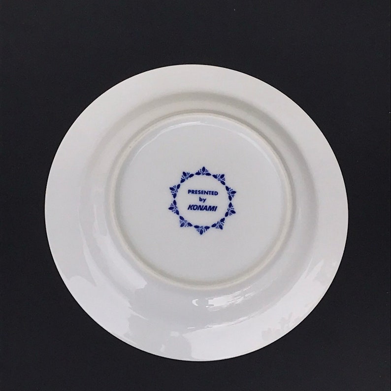 Op de afbeelding: Een wit keramiek bord met een blauw en wit ontwerp. Het bord heeft een blauwe cirkel met de tekst "PRESENTED by KONAMI" in het midden.