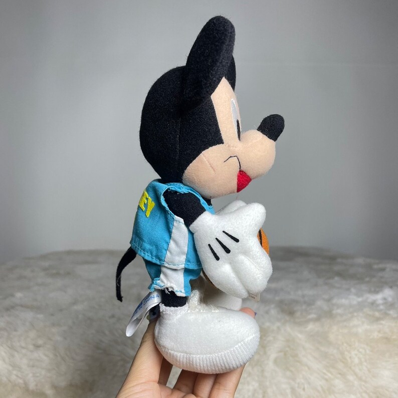 K&ouml;nnte beinhalten: Ein Pl&uuml;sch-Mickey-Mouse-Spielzeug, das eine blaue und wei&szlig;e Basketballuniform tr&auml;gt und einen Basketball h&auml;lt. Das Spielzeug hat eine schwarze Nase und rote Wangen.