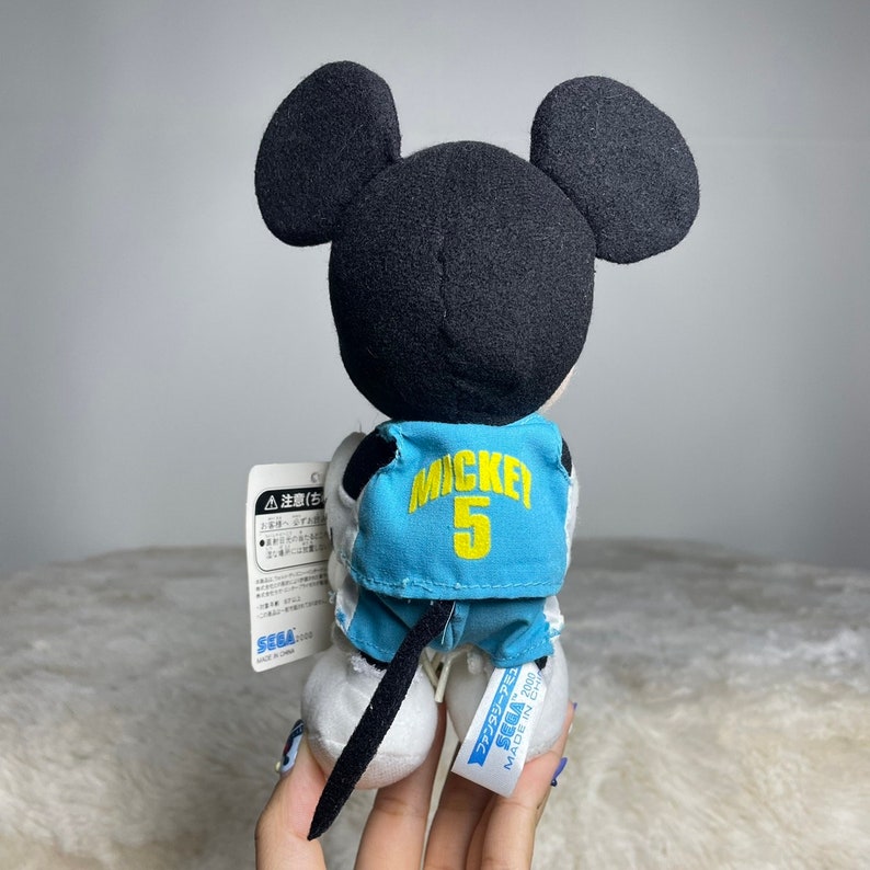 K&ouml;nnte beinhalten: Ein Pl&uuml;sch-Mickey Mouse, der ein blaues Basketballtrikot mit dem Wort "Mickey" und der Nummer "5" auf dem R&uuml;cken tr&auml;gt. Das Pl&uuml;schtier h&auml;lt einen Basketball und hat ein Etikett an seinem Bein, auf dem "Sega Fantasy Star Online 2001 Made in China" steht.
