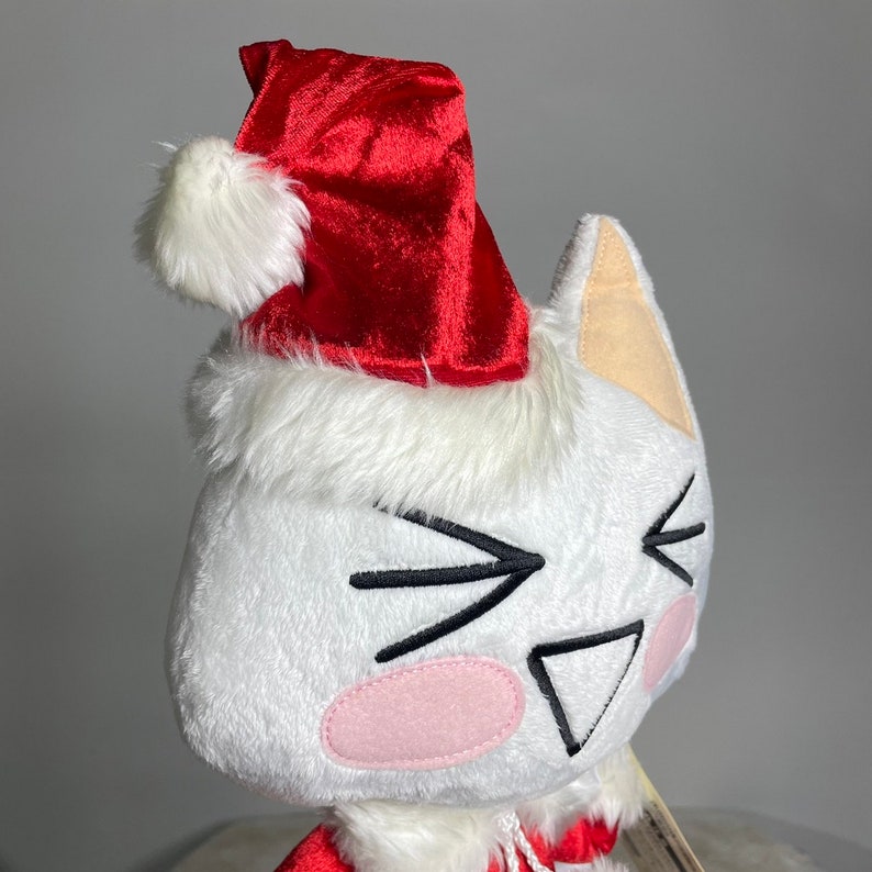May include: White plush toy cat wearing a red velvet Santa hat with a white fur trim. The cat has a black embroidered face with a pink nose and a mischievous expression.