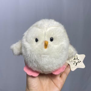 Könnte beinhalten: Eine weiße Plüsch-Eule mit rosa Füßen und einem gelben Schnabel. Die Eule sitzt auf einer Hand und hat ein kleines Etikett, auf dem in japanischen Schriftzeichen "Ofun" steht.