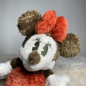 May include: A plush toy of a cartoon mouse with brown and white fur. The mouse has a large red bow and embroidered eyes. The toy is sitting on a white fluffy surface.