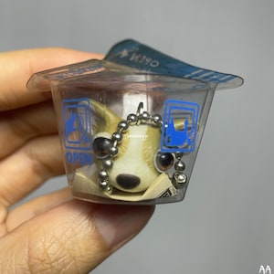 May include: A small, brown and white dog figurine keychain in a clear plastic container with a blue and white label that says "OPEN".
