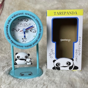 以下が含まれることがあります： パンダのデザインが施された、水色のTarepanda卓上時計。時計の文字盤には日本語のテキストとパンダのイラストが描かれています。時計はパンダのフィギュアが付いたスタンドで支えられています。お揃いの箱が右側にあります。