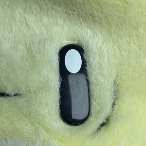 May include: A black and white button with a white circle in the center. The button is attached to a plush, light yellow fabric.