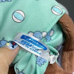 Może przedstawiać: Pluszowa zabawka w jasnozielonym kolorze z brązową twarzą i białą metką z napisem "Fun★Fan Amuse Prize Collection SEGA&reg; 2005 Made in China". Zabawka ma na sobie jasnozielony i niebieski str&oacute;j w wzory z białymi kulkami.