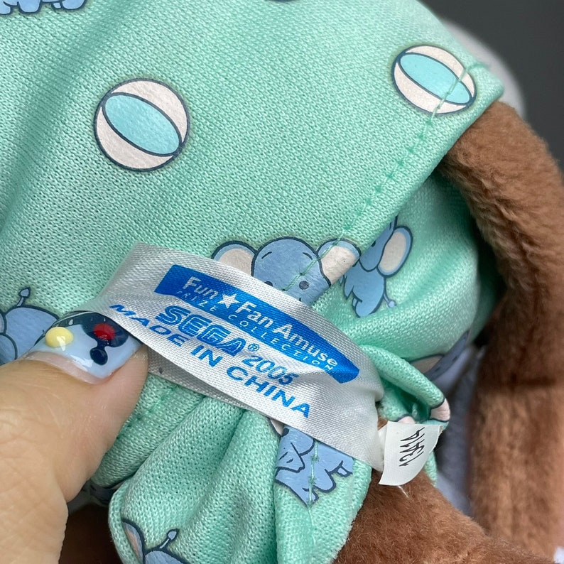Może przedstawiać: Pluszowa zabawka w jasnozielonym kolorze z brązową twarzą i białą metką z napisem "Fun★Fan Amuse Prize Collection SEGA&reg; 2005 Made in China". Zabawka ma na sobie jasnozielony i niebieski str&oacute;j w wzory z białymi kulkami.