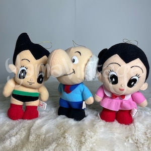 May include: Three plush toys from the Astro Boy anime series. The toys are Astro Boy, Dr. Tenma, and Uran. Astro Boy is wearing a green and black outfit, Dr. Tenma is wearing a blue shirt and black pants, and Uran is wearing a pink dress.